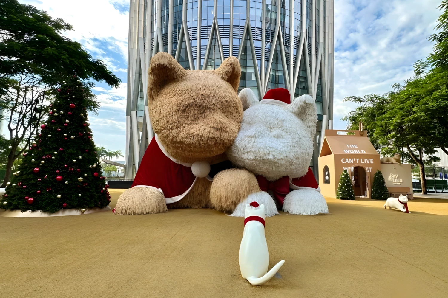 Giant plush yellow and white cat outdoor art sculpture with modern glass skyscraper background, beside Christmas trees and a giant dog house, with a small white cat statue on the plaza.