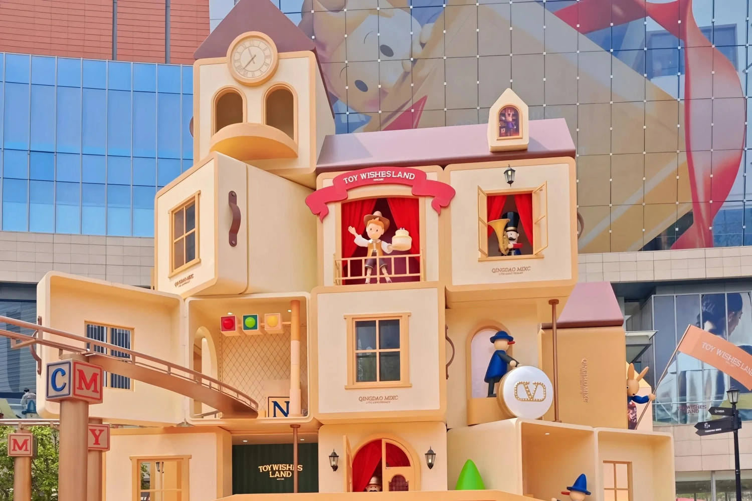 Front panoramic view of the "Toy Wishesland" mall installation in Qingdao, featuring block-style wooden toy houses, a clock tower, and musical character figures.