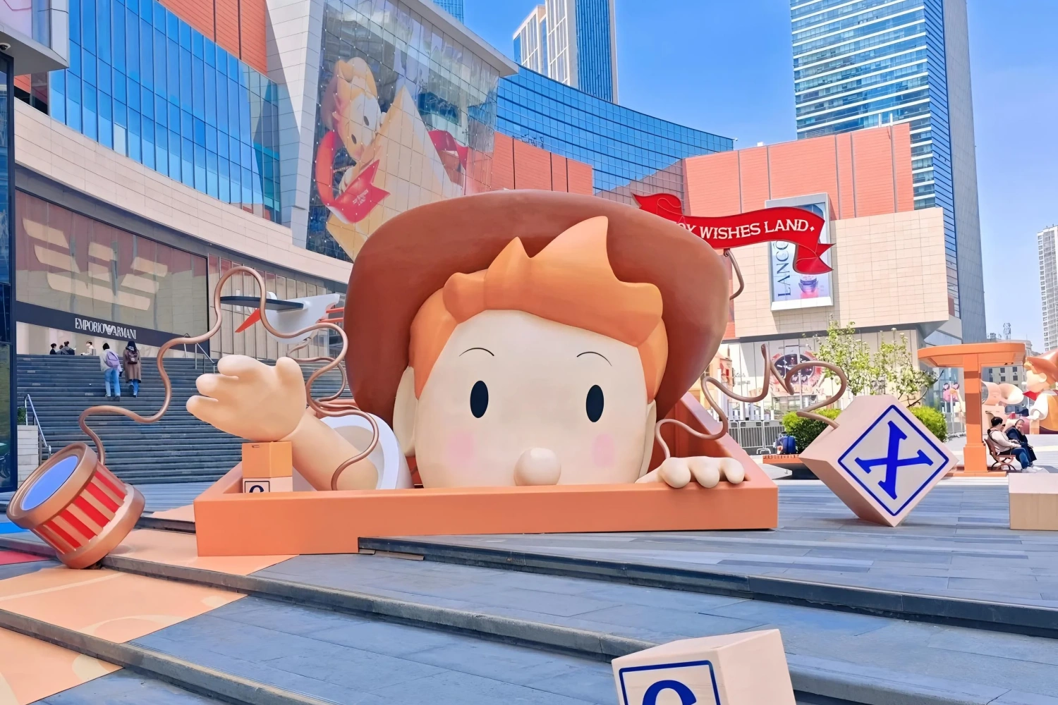 A giant cartoon boy head sculpture sitting on outdoor mall stairs, emerging from a yellow wooden frame, surrounded by wind-up toy elements.