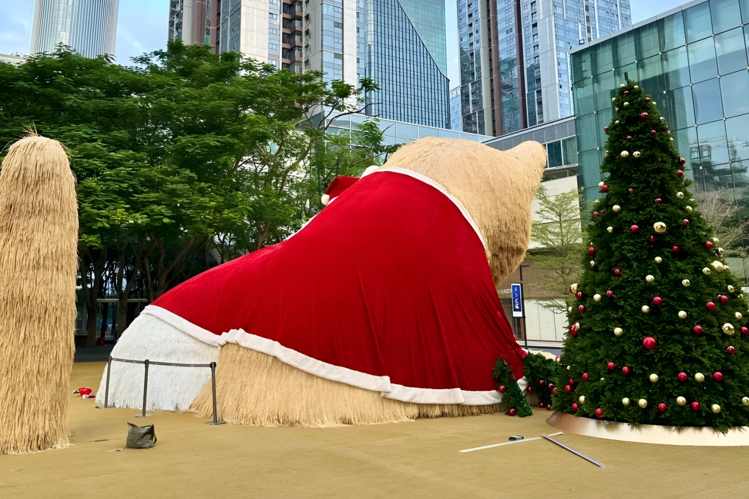 Back view of a giant plush cat sculpture wearing a red Christmas cape, situated in a city plaza, with a tall Christmas tree decorated with ornaments on the right.