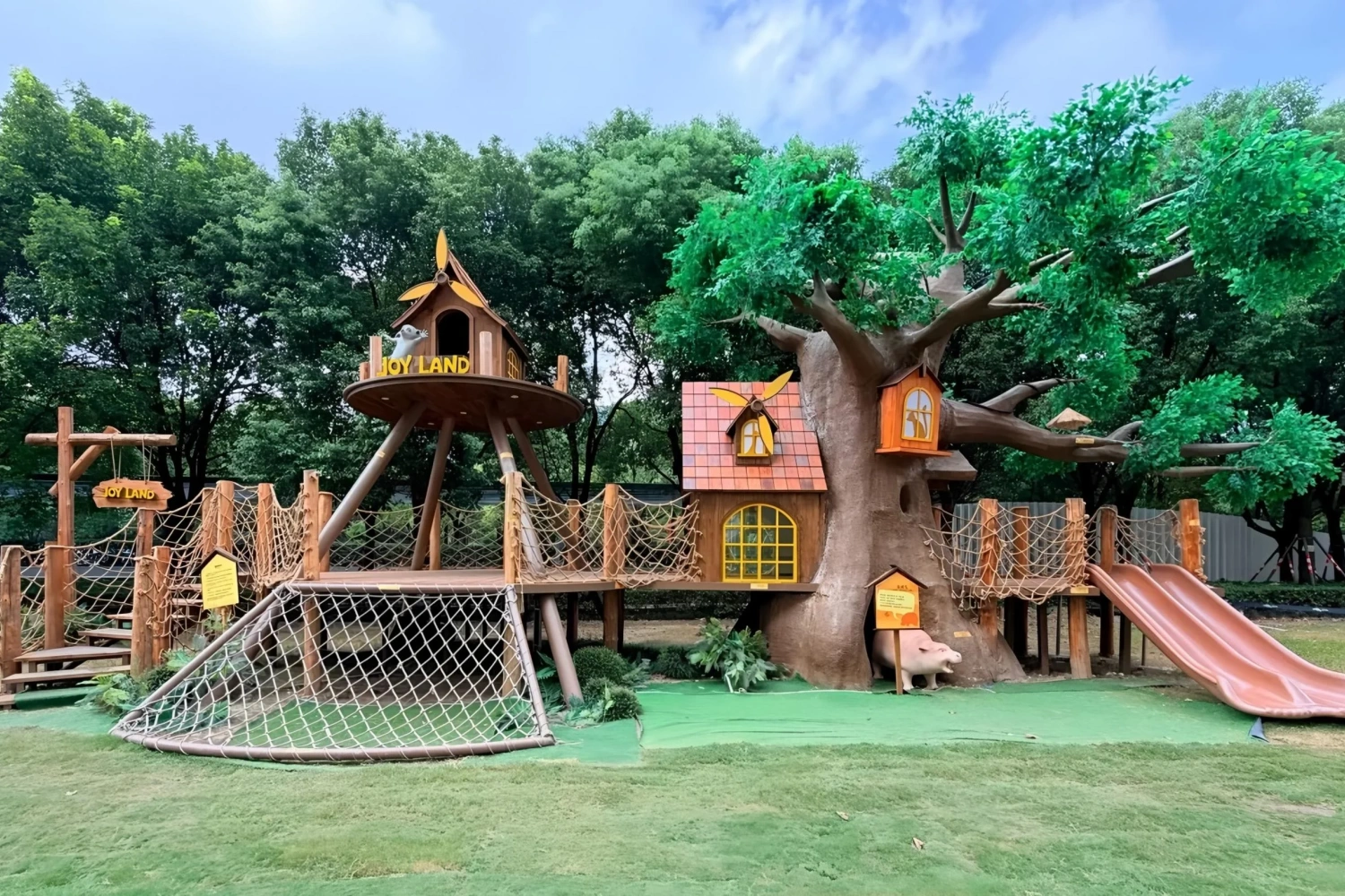 Completed large-scale forest theme landscape, featuring a multi-tiered play structure with rope bridges, a slide, and a "JOY LAND" treehouse, perfectly situated on an outdoor lawn.