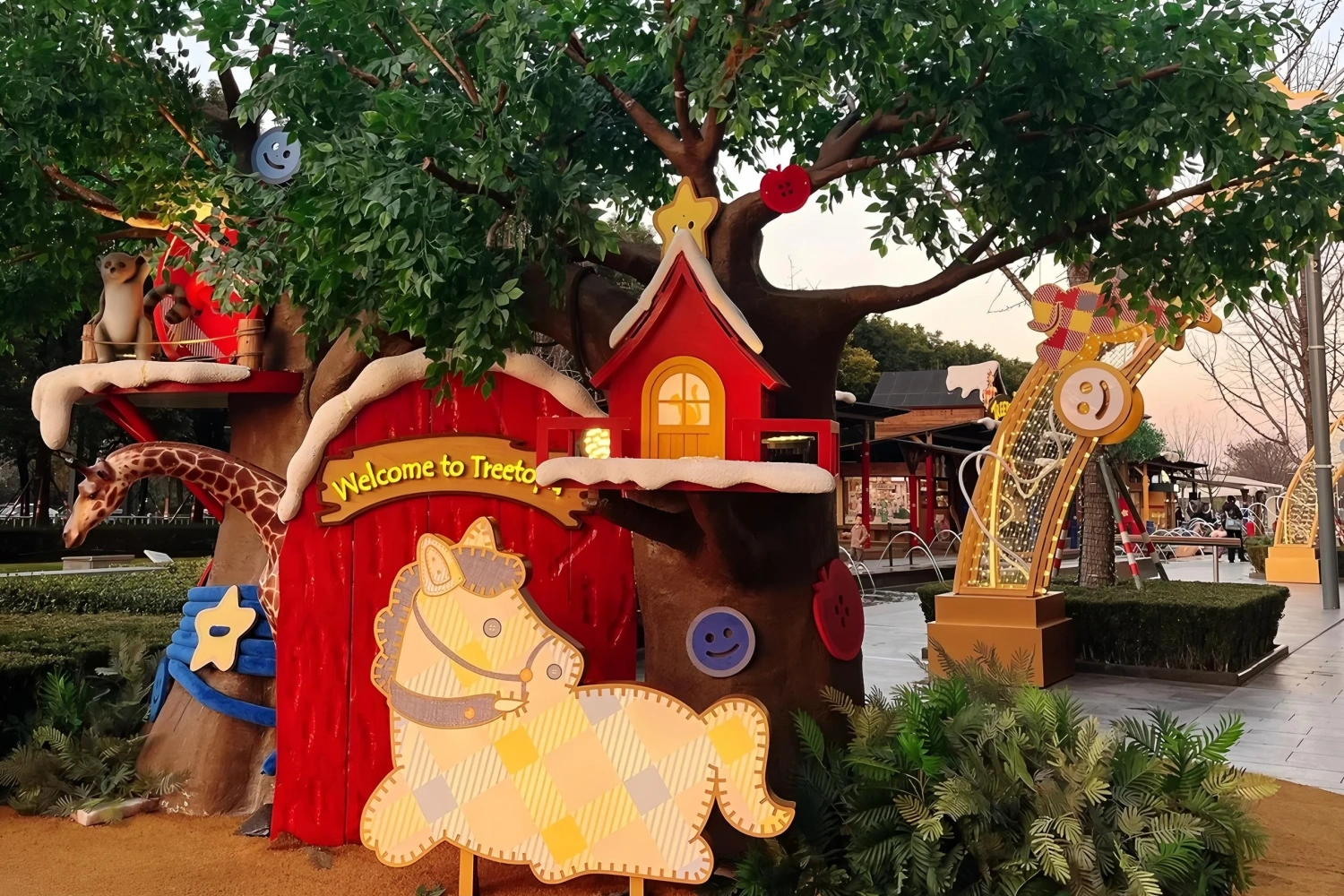 Close-up of forest theme mall decoration, featuring a red treehouse with yellow windows and snow-trimmed edges, alongside cute giraffe and zebra plush models, creating a fairy-tale interactive photo spot.