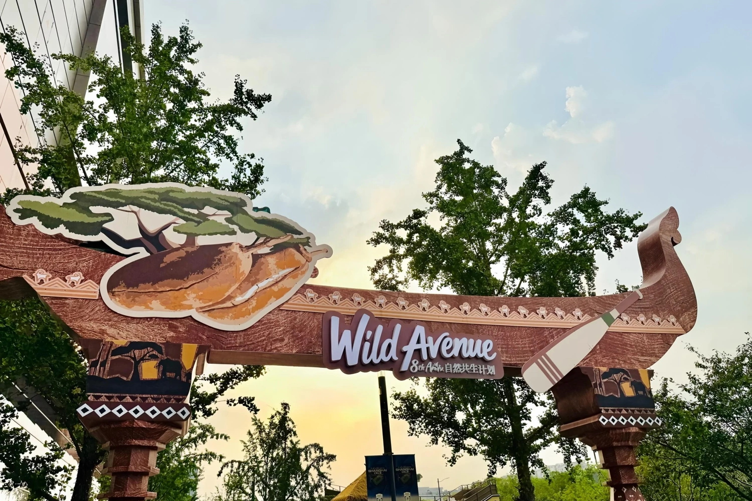 "Wild Avenue Nature Symbiosis Project" themed wooden archway at a commercial plaza. The top features a unique tree and terrain shaped display board, with a paddle decoration at the bottom right, fitting the nature exploration theme.