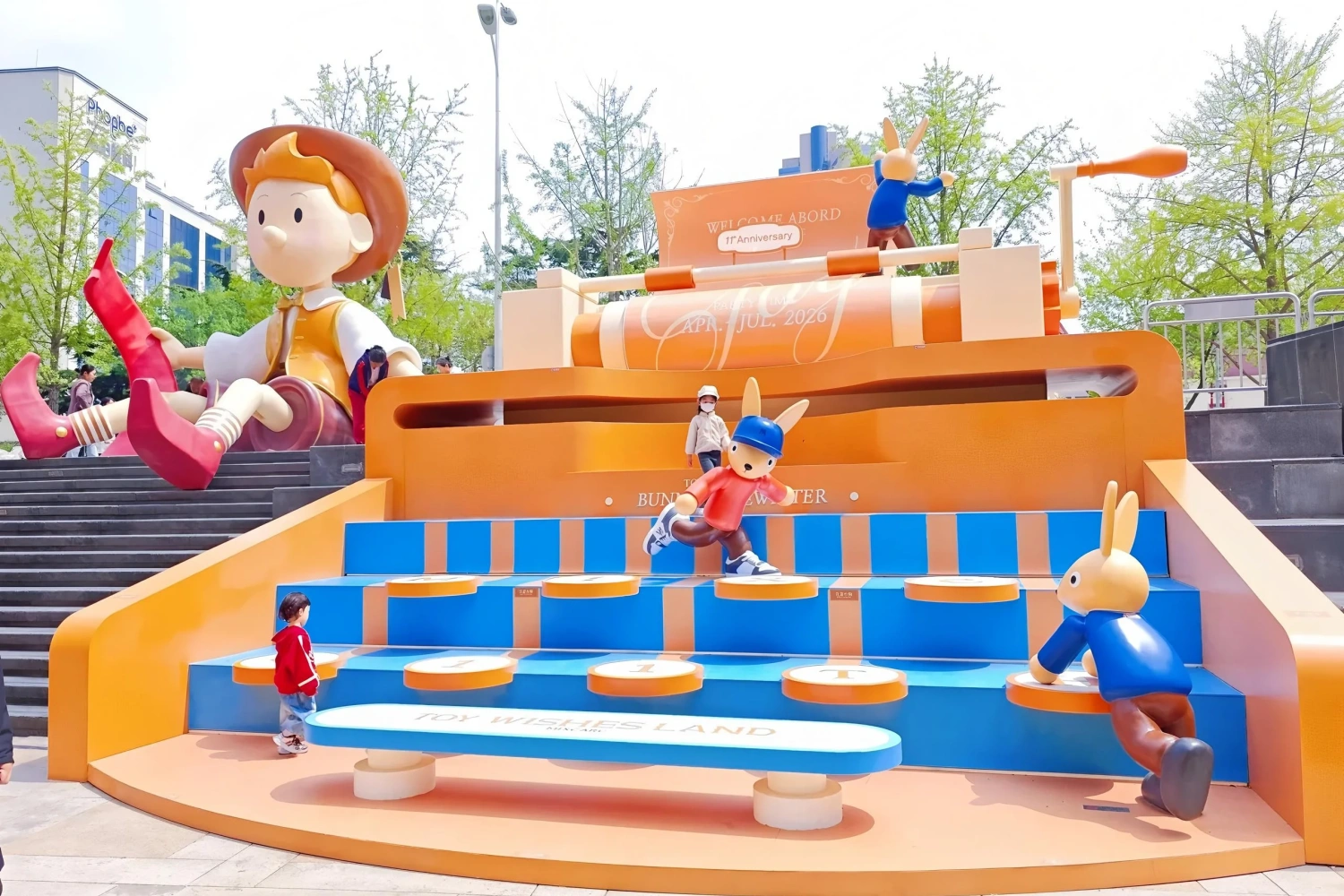 The complete interactive stair installation of "Toy Wishesland" in front of the mall plaza, featuring a giant boy sculpture, blue steps, and several cartoon bunny characters.