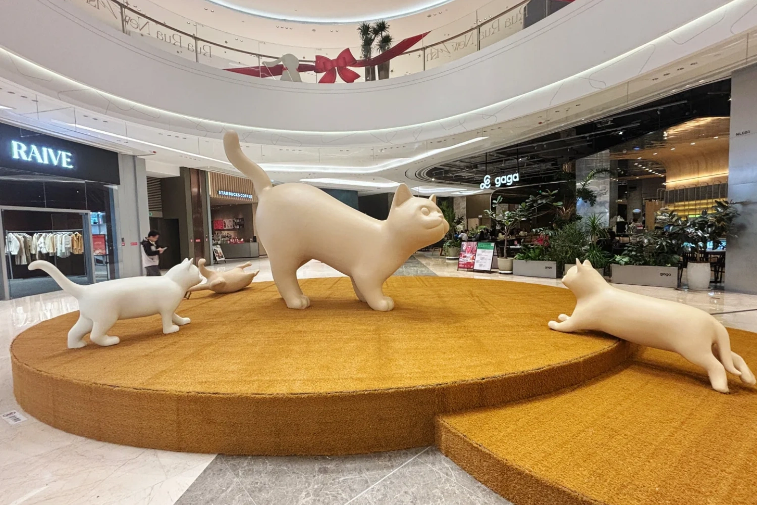 Panoramic view of the cat art installation in the mall atrium, featuring white and yellow cat sculptures in various poses on a large yellow circular carpet, creating a lively atmosphere.