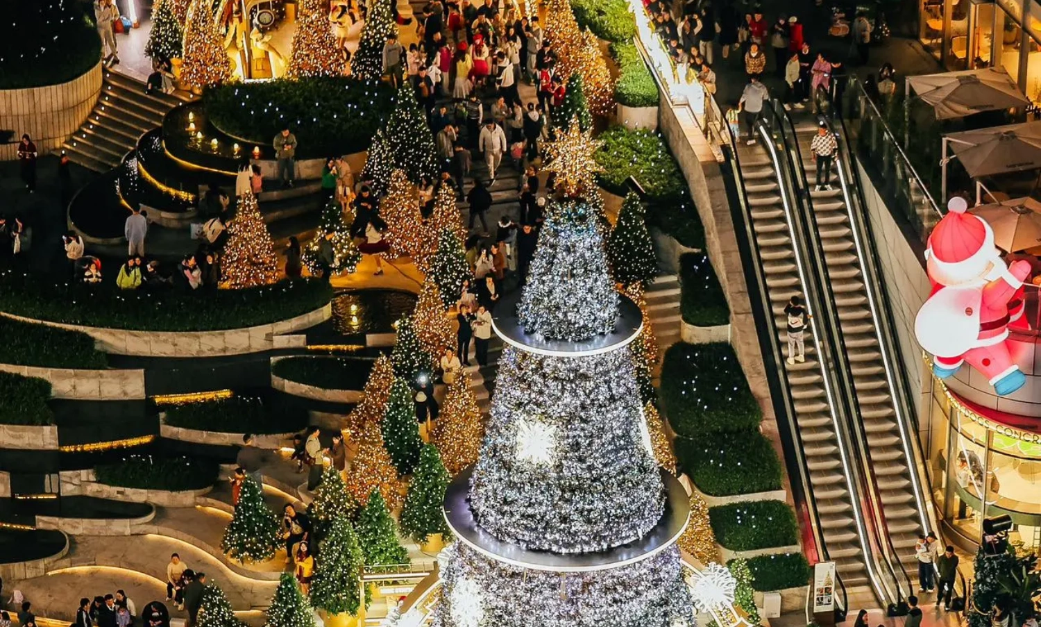 The MIXC mall's large-scale outdoor Christmas installation project, with its stunning visual volume and exquisite details, has become an influential and eye-catching winter landscape in the city. From the perspective of professional factory custom fabrication and on-site execution, this is a benchmark case integrating irregular structural processing, complex lighting engineering, and precise on-site construction solutions. Below, we provide an in-depth analysis of the project's entire execution process across five core dimensions:
1. Structural Safety: Wind Load Challenges of Ultra-High Tower Structures
The massive cone-shaped main tree stands in the center of the commercial plaza, featuring a layered ring structure design topped with a shining star decoration. As a large-scale customized outdoor commercial visual merchandising (VM) project, the primary challenge was passing the structural safety test in winter's strong wind environments. Once the main tree is heavily laden with dense ornaments and string lights, the windward resistance becomes immense, and the overall center of gravity is quite high.
During the prop fabrication phase, the factory utilized high-strength hot-dip galvanized steel for the core load-bearing skeleton, and structural engineers conducted rigorous wind load mechanics calculations. The base system employed a concealed counterweight foundation, while the upper tower body utilized modular flange bolting technology. This solid internal-to-external support ensured the absolute safety of the entire Christmas installation under extreme outdoor weather conditions.
2. Lighting & Craftsmanship Aesthetics: Seamless Arrangement of a Million-Level Starlight Matrix
The main body of the installation is covered with dense, waterfall-like cool white LED string lights, interspersed with large glowing snowflake motifs. On the other side, the "MIXC STATION" area features warm, golden-yellow lighting with a retro glow, complete with a delicate British-style double-sided clock. The outdoor lighting engineering not only requires the wiring to achieve an IP65 or higher waterproof rating, but more challenging is how to perfectly conceal the wiring among thousands of light points to maintain surface smoothness and craftsmanship aesthetics during the day when the lights are off.
The factory team customized a high-density, flame-retardant, light-transmitting mesh as the skeleton backing, and all main cables utilized an embedded, hidden wiring process. For color selection, the color temperature difference between the cool white and warm yellow lights was strictly compared. Through precise zoning, a strong visual contrast was created between the crisp snow-crystal texture and the warm, vintage atmosphere of the passageway.
3. Complete Pre-Assembly: 1:1 Precise Restoration of Irregular Components
Not only does the main tree require seamless splicing, but the smoothly curved golden archway supports and the "MIXC STATION" hanging plaque with its custom baked-enamel finish also demand extremely high dimensional accuracy. Multi-curved metal pipes are highly susceptible to stress deformation during bending and welding. Any tolerance discrepancies during on-site assembly would severely compromise the final execution of the VM design.
We comprehensively implemented a 1:1 complete pre-assembly protocol prior to factory dispatch. All curved metal structures and luminous rings underwent trial assembly in our extra-large factory workshop. The quality inspection department verified every screw hole and joint flatness one by one, proactively preventing potential alignment errors during on-site construction to ensure every module interlocked smoothly.
4. On-Site Installation & Detailing: Extreme Micro-Operations in Complex Terrain
The installation site featured a complex landscape with multi-level steps, water feature edges, and dense greenery; additionally, a giant inflatable Santa Claus was suspended from a surrounding wall. Mall installations are typically restricted to a very short time window at night after closing. Furthermore, the operating space for large hoisting equipment was limited on the multi-tiered plaza, and strict measures were required to protect the premium stone flooring from damage.
The construction team developed a highly modular, "building-block" style night operation plan. All load-bearing bases were pre-laid with custom flexible non-slip protective mats. Through the seamless coordination of heavy cranes and aerial work platforms, components were hoisted and assembled layer by layer from bottom to top according to their pre-assigned numbers. For edge detailing, custom metal skirting boards and artificial pine needles were cleverly utilized as transitional covers at the bottom edge, achieving a flawless, high-quality delivery(4)